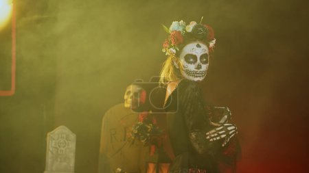 Photo for Glamorous woman with sugar skull make up and flowers headband, dressed in goddess of death costume as santa muerte. Dios de los muertos art to celebrate mexican tradition in studio. Handheld shot. - Royalty Free Image