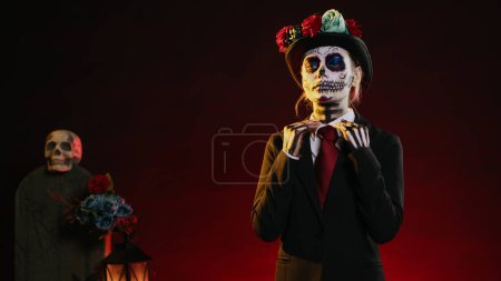 Photo for Goddess of dead with suit and hat looking creepy wearing festival skull make up or body art paint, celebrating dios de los muertos holiday. Acting like la cavalera catrina. Handheld shot. - Royalty Free Image