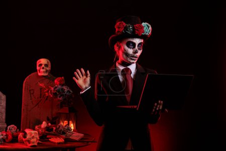 Photo for Scary woman using laptop on dios de los muertos mexican holiday, celebrating day of the dead with santa muerte costume. Wearing goddess of death skull body art, looking at wireless pc. - Royalty Free Image