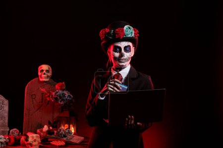Photo for Young adult doing online shopping with skull make up, using credit card on laptop to make payment. Wearing santa muerte lady of death costume to celebrate mexican halloween holiday. - Royalty Free Image