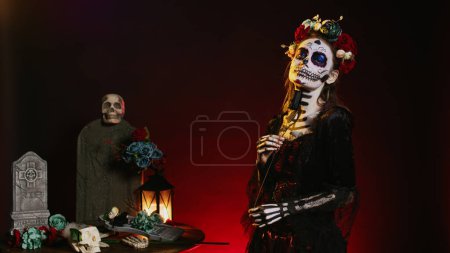 Foto de Modelo femenina vestida con arte corporal de santa muerte en estudio, actuando glamorosa el día de la celebración de Halloween mexicana muerta. Arte corporal de cráneo ritual, posando con rosas negras y corona de flores. - Imagen libre de derechos