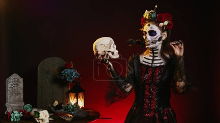 Téléchargez les photos : Femme effrayante agissant effrayant avec le crâne à la main, regardant le crâne d'Halloween. Portant un maquillage effrayant ressemblant à Santa Muerte avec couronne de fleurs et art corporel sur le rituel saint mexicain. - en image libre de droit