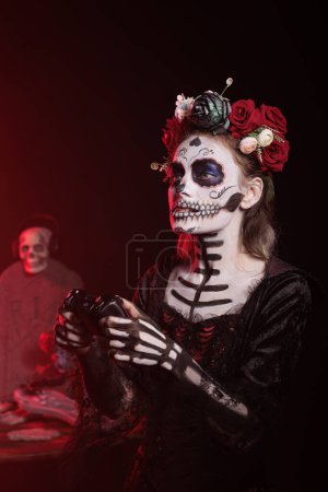 Foto de Lady of death jugando videojuegos en línea con joystick en la consola, usando maquillaje de cráneo y disfraz tradicional de santa muerte. Mujer disfrutando de la competencia de juego en el día de la muerte. - Imagen libre de derechos