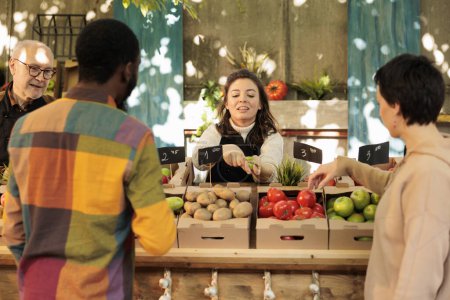 Téléchargez les photos : Femme souriante gaie offrant au client d'essayer un petit morceau de pomme tout en vendant des fruits et légumes biologiques frais au marché fermier local, accent sélectif. Dégustation pendant les courses - en image libre de droit