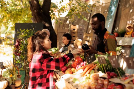 Téléchargez les photos : Vendeur afro-américain debout derrière étal vendant des légumes frais biologiques de saison à la clientèle féminine au marché fermier local. Jeune femme achetant des produits naturels maison - en image libre de droit