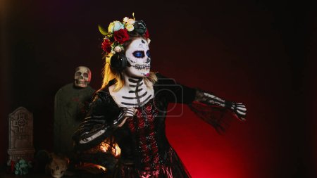 Photo for La cavalera catrina listening to music on audio headset, using headphones to enjoy mp3 song over black background. Creepy horror woman using headphones on holy dios de los muertos tradition. - Royalty Free Image