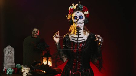 Photo for Goddess of dead enjoying music on headphones, wearing skull make up and santa muerte costume in studio. Beautiful person listening to song on audio headset, celebrating day of the dead. - Royalty Free Image