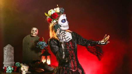 Photo for Young scary woman talking on smartphone call, wearing halloween make up and flowers crown in studio. La cavalera catrina using smartphone to chat on dios de los muertos mexican holiday. - Royalty Free Image