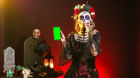 Photo for Creepy female model holding greenscreen on smartphone, using mobile phone with isolated mockup template. Woman dressed as santa muerte showing blank chroma key copyspace display. - Royalty Free Image