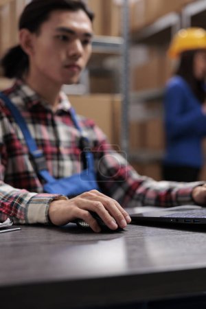 Photo for Young asian logistics manager analyzing goods supply on laptop. Warehouse worker doing inventory management on computer while sitting at desk with close up selective focus on hand - Royalty Free Image