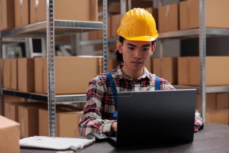 Photo for Young asian man warehouse operator managing delivery on laptop while working at storage room desk. Storehouse manager in yellow helmet analyzing products logistics on computer - Royalty Free Image