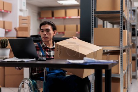 Asian wheelchair user managing parcel delivery in postal service warehouse. Storehouse shipment operator with disability holding cardboard box with customer order and planning shipping route on laptop
