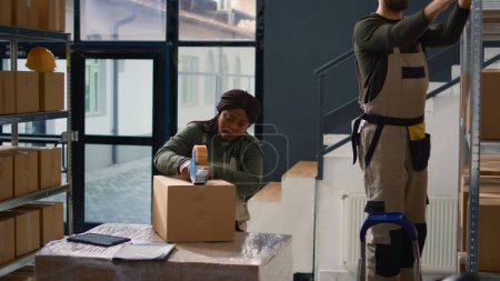 Photo for Dolly out shot of head of operations in warehouse supervising trainee sealing cardboard box parcels. Retail depository personnel properly securing goods to avoid damages during shipping - Royalty Free Image