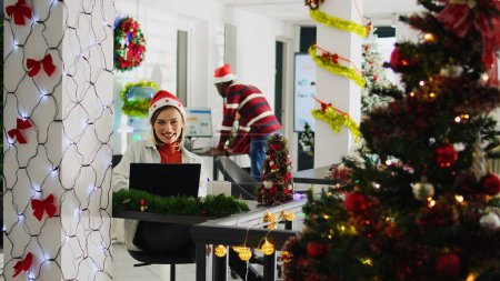 Téléchargez les photos : Dolly out portrait prise de vue d'un employé heureux travaillant sur la saison de Noël dans un bureau décoré. Travailleur joyeux résoudre les tâches au bureau dans divers lieux de travail décorés de Noël à côté de collègues - en image libre de droit
