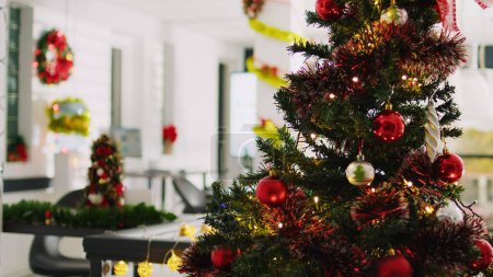 Photo for Jib up close up shot of beautifully ornate Christmas pine tree with empty office in blurry background. Xmas tree adorn with red garlands and baubles in festive workspace during winter holiday season - Royalty Free Image