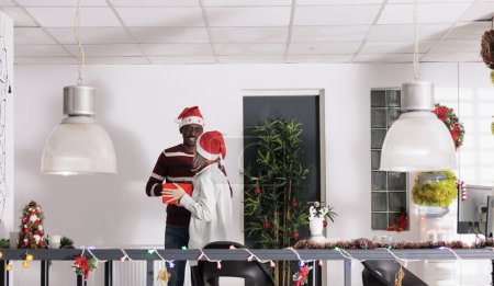 Foto de El empleado feliz recibe el regalo secreto de Papá Noel del compañero de trabajo en la temporada navideña en la oficina decorada. Sonriente trabajador afroamericano recibe regalo de Navidad en vacaciones de invierno festivas - Imagen libre de derechos