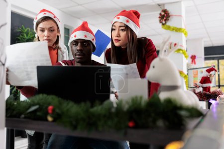Photo for Employees comparing business analytics data charts in festive decorated office. Multiethnical coworkers solving tasks in Christmas ornate workspace during winter holiday season - Royalty Free Image