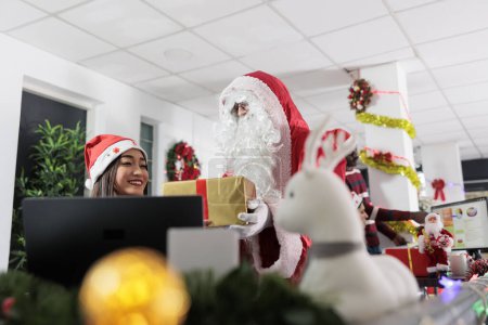 Foto de Gerente vestido como Santa Claus sorprendente empleado de BIPOC con regalo creativo de Navidad en el espacio de trabajo ornamentado festivo. Ejecutivo de gestión que ofrece caja de regalo para compañeros de trabajo durante la temporada navideña de Navidad - Imagen libre de derechos