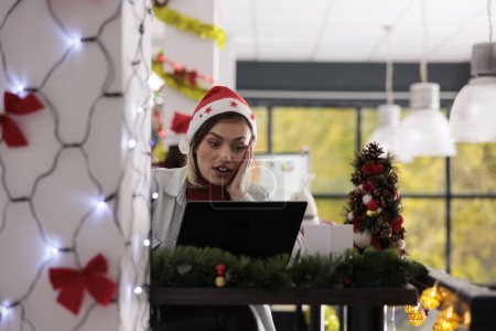 Photo for Businesswoman wearing Christmas hat in festive decorated office shocked by company quarterly finance revenue. Employee sitting at desk working in xmas adorn workspace during winter holiday season - Royalty Free Image