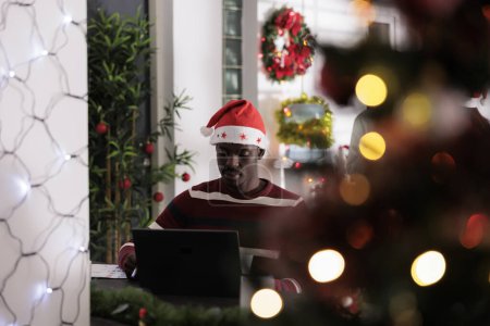 Téléchargez les photos : Un employé méticuleux portant des vêtements sur le thème de Noël s'est concentré sur la finition de la paperasserie pendant les vacances d'hiver. Expérimenté travailleur afro-américain résoudre des tâches dans Noël festive ornent bureau - en image libre de droit