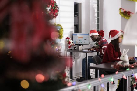 Photo for Focused african american businessman solving tasks on computer sitting at desk during holiday season. Meticulous employee wearing christmas hat, working in xmas decorated workplace office - Royalty Free Image