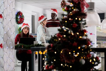 Photo for Excited company manager enjoying Christmas spirit mood, bringing package at work full of festive ornaments helped by employee. Business team leader adorning workplace with xmas decorations - Royalty Free Image