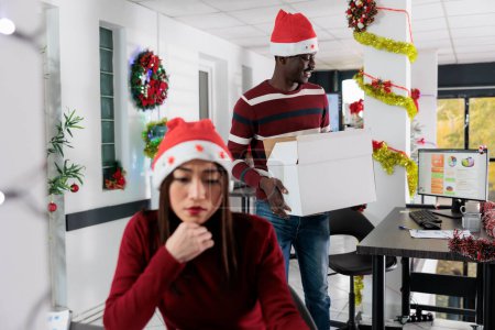 Téléchargez les photos : Un employé afro-américain emménage dans un nouveau bureau décoré pour Noël après avoir accepté un nouveau poste dans l'entreprise. Travailleur excité de commencer un nouvel emploi dans un espace de travail décoré et festif - en image libre de droit