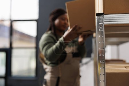 Photo for Selective focus of metallic shelves full with carton boxes, african american worker preparing customers order in storage room. Stockroom employee wearing industrial overall working at storehouse - Royalty Free Image