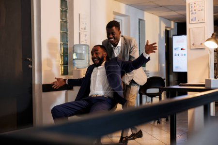 Téléchargez les photos : Travailleurs afro-américains ayant compétition de course sportive dans le bureau de démarrage moderne, prendre une pause du travail et avoir du plaisir. hommes d'affaires joyeux en costume formel chaise de bureau équitation tard dans la nuit - en image libre de droit