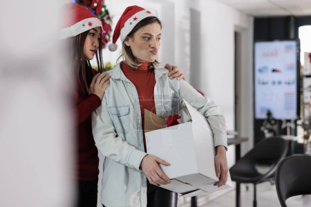 Photo for Asian employee upset by company manager leaving Christmas decorated office after deciding to quit. Team leader holding desk belongings box, bidding farewell to assistant before starting another job - Royalty Free Image