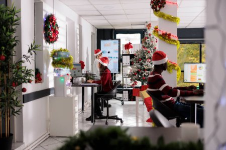 Photo for Multiethnical business team wearing christmas hats doing project tasks in festive workplace. Multicultural employees checking statistical graphs in decorated office during xmas season - Royalty Free Image