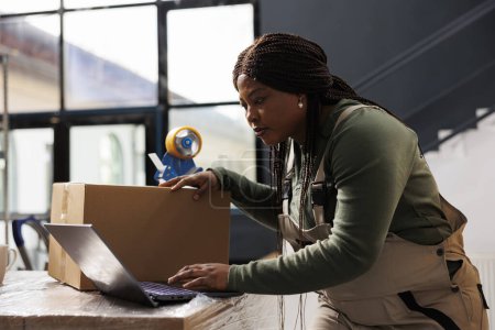 Photo for Warehouse manager checking online orders on laptop computer, analyzing shipping details while preparing packages. African american worker analyzing merchandise logistics in storage room - Royalty Free Image