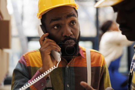 Photo for African american man talking on landline phone in industrial storehouse. Factory warehouse operator answering supervisor telephone call and listening to inventory management instructions - Royalty Free Image