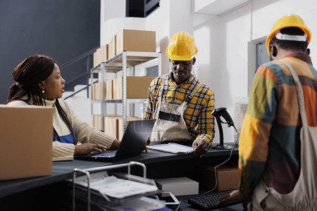 Photo for Shipment manager asking warehouse workers about merchandise storage system. African american storehouse employees talking and using inventory software on laptop at reception counter - Royalty Free Image