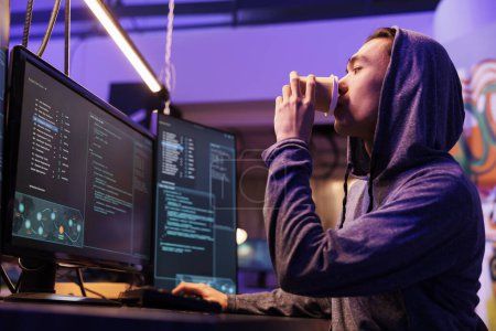 Photo for Hacker breaking website on computer while drinking coffee to go. Young asian man in hood cracking password using malicious software and getting illegal access to steal data - Royalty Free Image