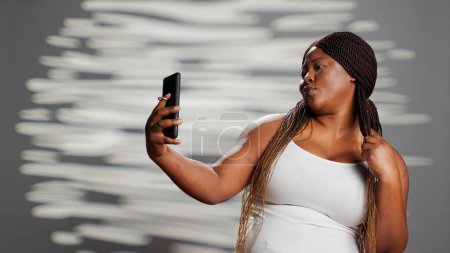 Téléchargez les photos : Femme afro-américaine se sentant confiante en prenant des photos, en utilisant un smartphone pour montrer l'amour de soi et la positivité du corps. Modèle féminin jouant dans le studio pour promouvoir la confiance en soi, la peau rayonnante. - en image libre de droit