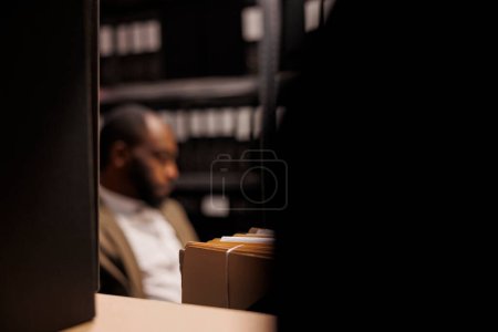 Photo for Shelf with criminal case files and policeman in dark police office. Blurred african american private investigator working overtime, solving crime at night time selective focus - Royalty Free Image