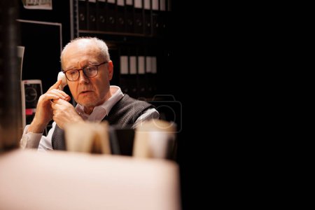 Photo for Elderly investigator discussing criminal case with remote officer using landline phone, working late at night in arhive room. Private detective analyzing criminology report. Investigation concept - Royalty Free Image