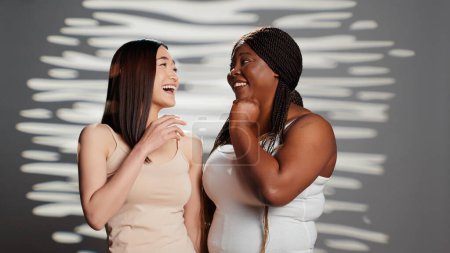 Photo for Curvy young adult telling secret to asian lady on camera, acting sensual and positive in studio. Diverse beauty models advertising body positivity and self confidence, having fun. - Royalty Free Image