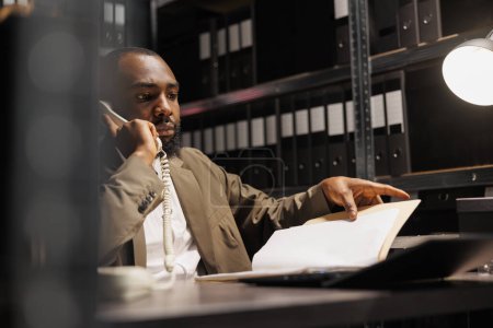 Photo for African american police investigator answering landline phone call in office at night time. Concerned detective discussing crime case insight while analyzing forensic report file - Royalty Free Image