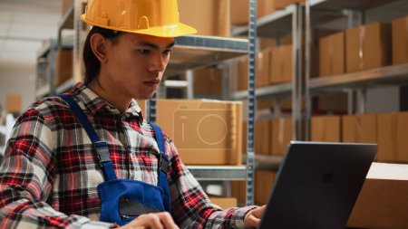 Photo for Asian man checking merchandise quality in warehouse, working on shipment and delivery with laptop. Young worker analyzing inventory for distribution management, small business concept. - Royalty Free Image