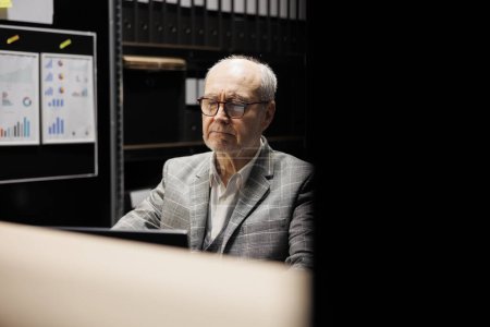 Photo for Senior businessman executive sitting at office desk, inspecting quarterly company results analysis data paperwork on laptop. Accountancy workplace filled with document folders on cabinet shelves - Royalty Free Image
