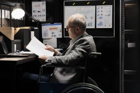 Photo for Professional accountant in wheelchair checking administrative paperwork in dark file room. Businessman in bureaucratic file cabinet office filled with invoice folders and flowcharts - Royalty Free Image