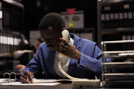 Foto de Gerente afroamericano hablando por teléfono fijo, discutiendo la investigación de gestión con el contable remoto en el almacén. Empresario trabajando hasta tarde por la noche en el informe de contabilidad - Imagen libre de derechos