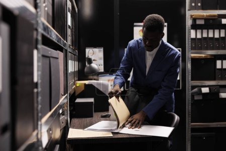 Foto de Gerente afroamericano recogiendo sus cosas, preparándose para dejar el trabajo tarde en la noche después de trabajar en el informe de contabilidad en la sala de archivos. Empleado en busca de registro de burocracia en depositario corporativo - Imagen libre de derechos
