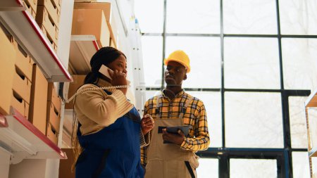 Photo for Team of people planning cargo shipment in warehouse, woman answering phone call in depot. Male employee checking list of products while manager talks on landline phone. Handheld shot. - Royalty Free Image