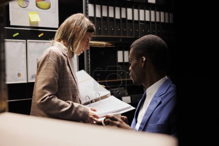 Photo for Diverse depository employees analyzing bureaucracy record, checking administrative documents late at night in storage room. Bookkeepers discussing accountancy report, discovering management files - Royalty Free Image