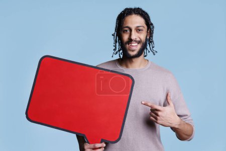 Photo for Smiling arab man pointing with finger at red speech bubble with copy space portrait. Cheerful happy person holding empty banner and advertising product while looking at camera - Royalty Free Image