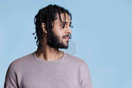 Photo for Arab man with neutral facial expression looking to left side. Young adult arabian handsome model with black braided hair posing for profile studio shot on blue background - Royalty Free Image