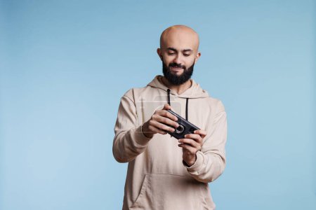Photo for Young arab man playing mobile game, using entertaining application software. Concentrated gamer enjoying videogame on smartphone while standing in studio on blue background - Royalty Free Image